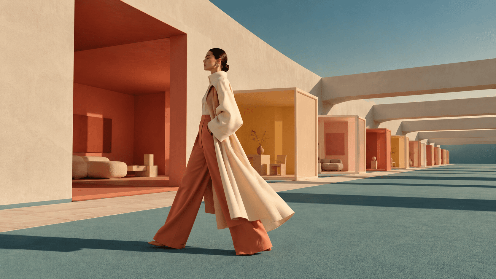 A woman in a flowing cream coat and wide rust-colored trousers walks through a minimalist architectural corridor.